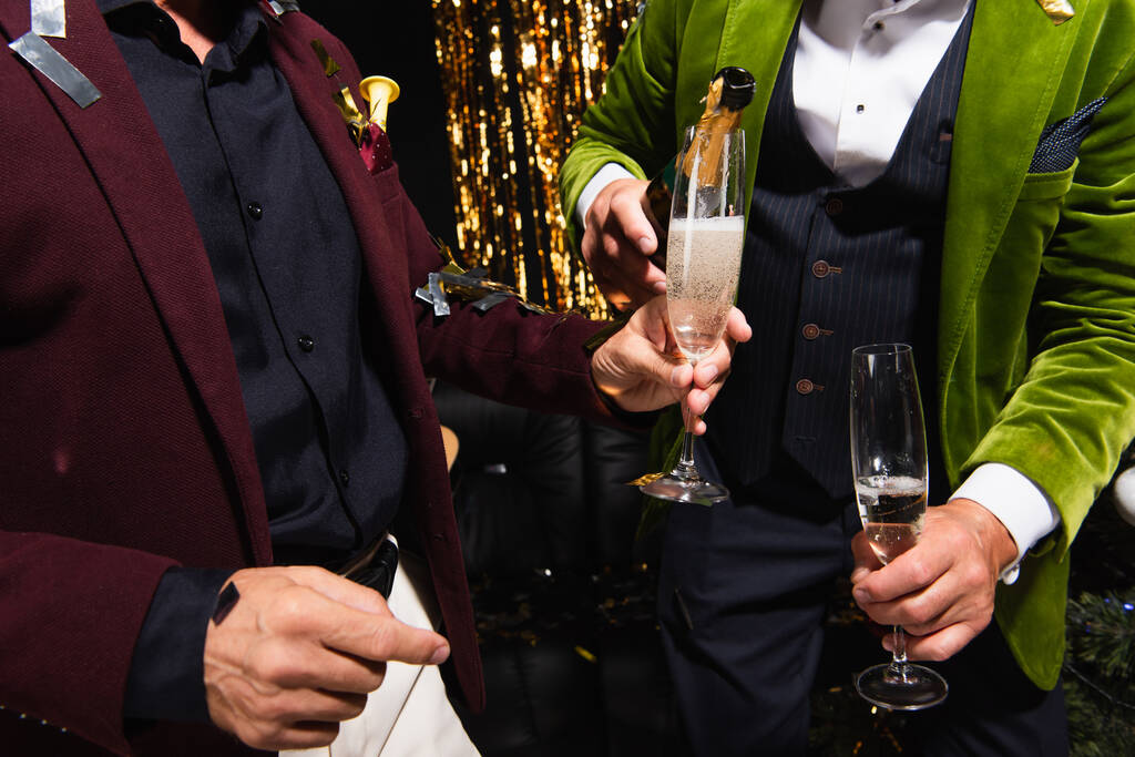 Vista ritagliata dell'uomo che tiene champagne vicino all'amico con bottiglia e coriandoli durante la festa su sfondo nero  - Foto, immagini