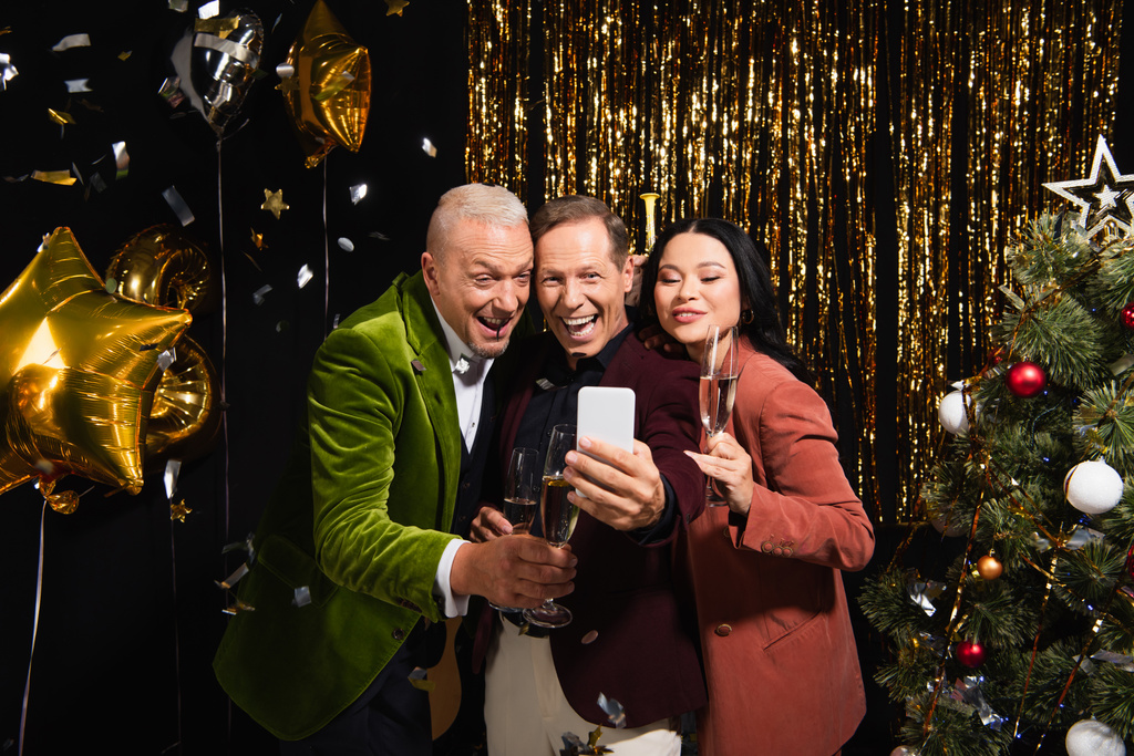Hymyilevä monikansallisia ystäviä samppanjaa ottaen selfie älypuhelimeen lähellä joulukuusi ja ilmapalloja mustalla taustalla  - Valokuva, kuva