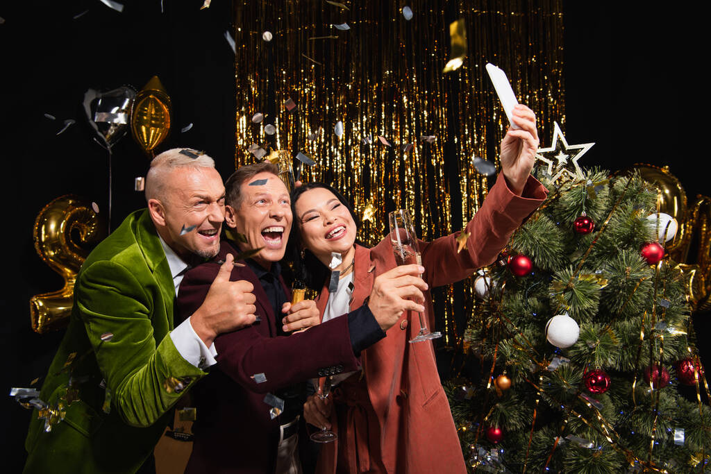 Happy interracial friends with champagne taking selfie on smartphone near christmas tree and confetti on black background  - Photo, Image
