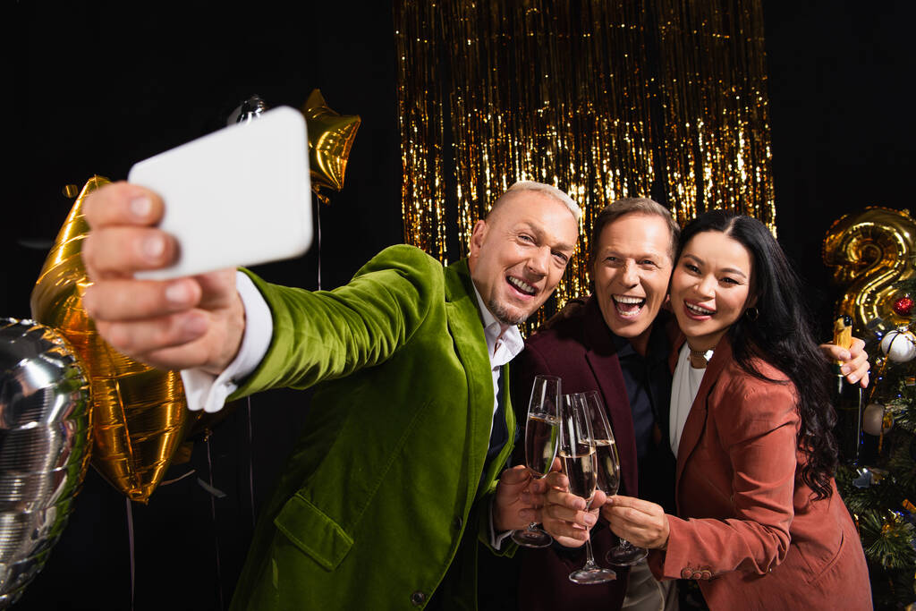Усміхнені багатоетнічні друзі з шампанським беруть селфі на розмитий смартфон під час новорічної вечірки на чорному тлі
  - Фото, зображення
