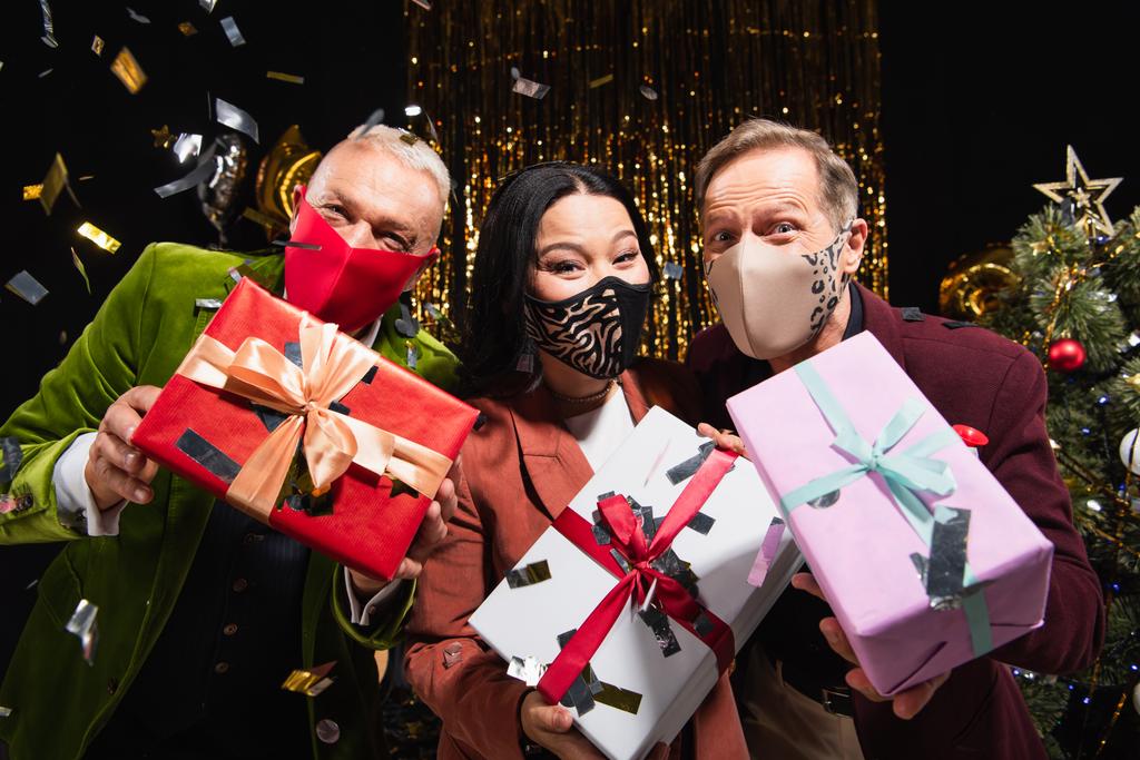 Multietnické přátelé v ochranných maskách držení dárky pod konfety během nového roku party na černém pozadí  - Fotografie, Obrázek