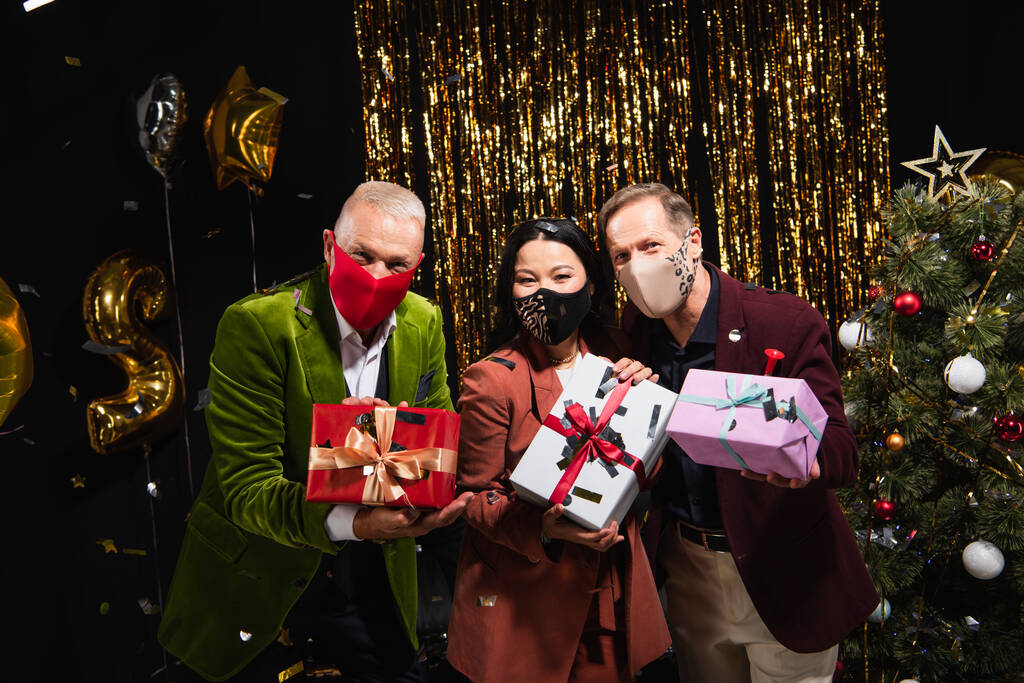 Reife interrassische Freunde in Schutzmasken mit Geschenken in der Nähe von Weihnachtsbaum und Luftballons auf schwarzem Hintergrund  - Foto, Bild
