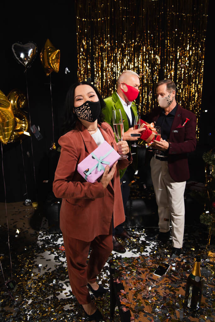 Mujer asiática en máscara protectora celebración de champán y presente cerca de amigos durante la fiesta de año nuevo sobre fondo negro  - Foto, Imagen