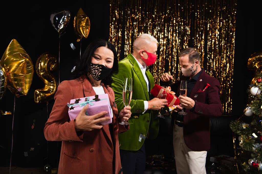 Koruyucu maskeli olgun bir Asyalı kadın elinde şampanya tutuyor ve yeni yıl partisinde siyah arka planda arkadaşlarının yanında.  - Fotoğraf, Görsel