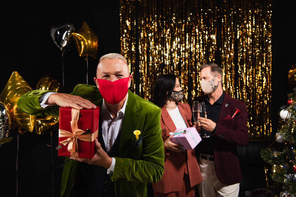Mature man in formal wear and protective mask holding present near interracial friends and festive decor on black background  - Photo, Image