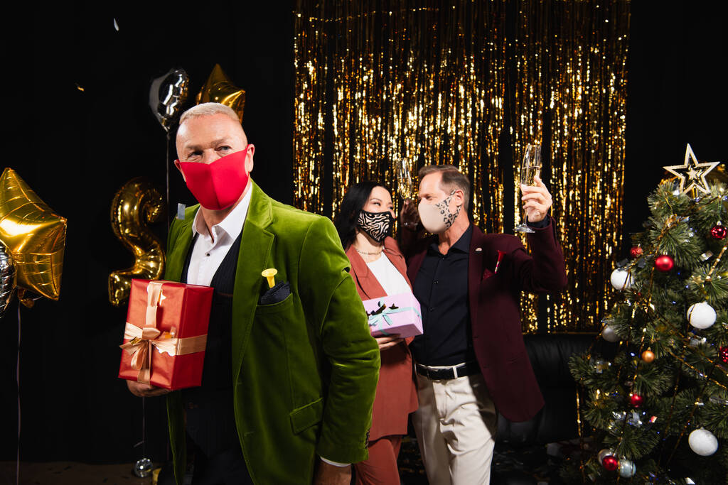 Mature man in protective mask holding gift near multiethnic friends with champagne and christmas tree on black background  - Photo, Image