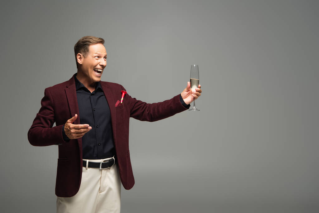 Hombre positivo en ropa formal apuntando a la copa de champán aislado en gris  - Foto, imagen
