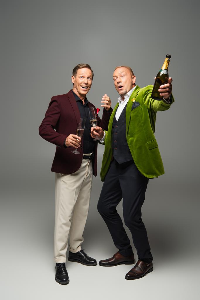 opgewonden volwassen mannen met champagne en fles kijken naar camera op grijze achtergrond  - Foto, afbeelding