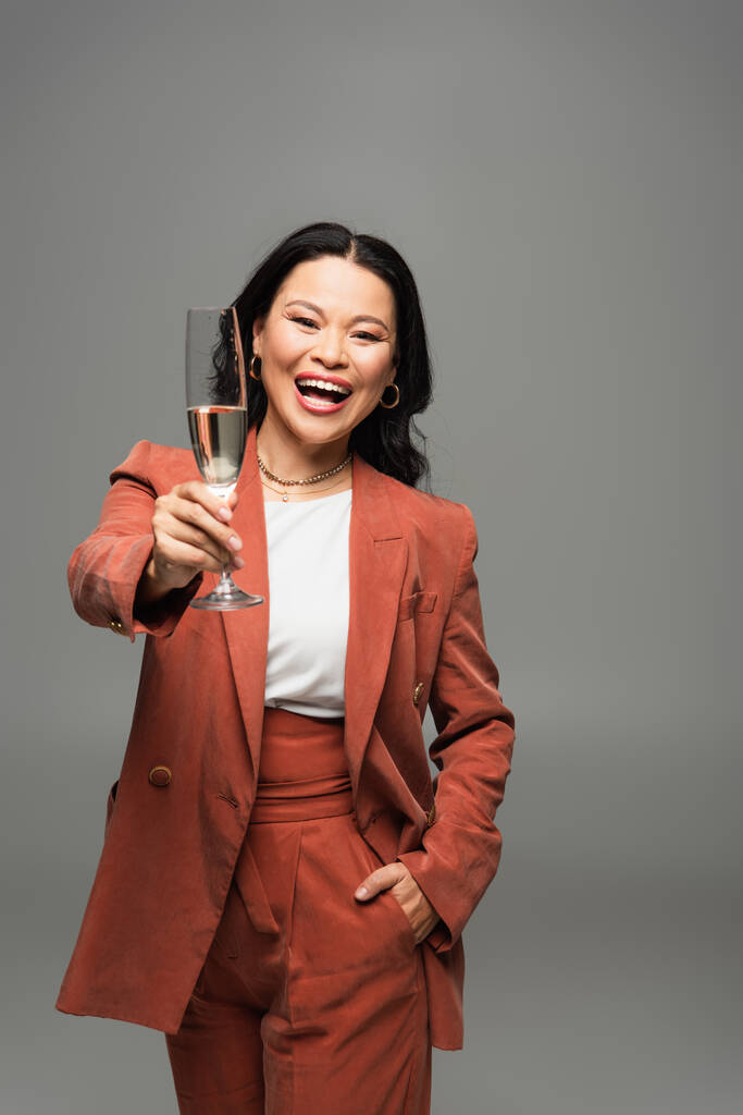 Femme asiatique positive en veste en terre cuite tenant un verre de champagne isolé sur gris  - Photo, image