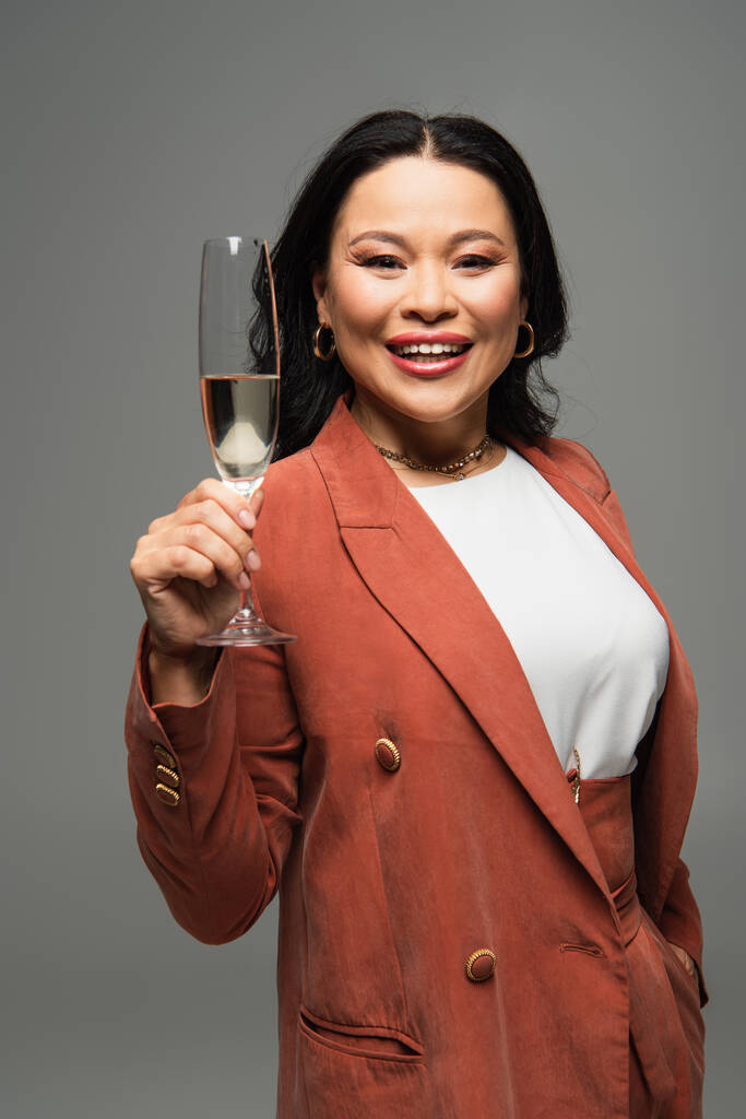 Mature asian woman holding glass of champagne and smiling at camera isolated on grey  - Photo, Image