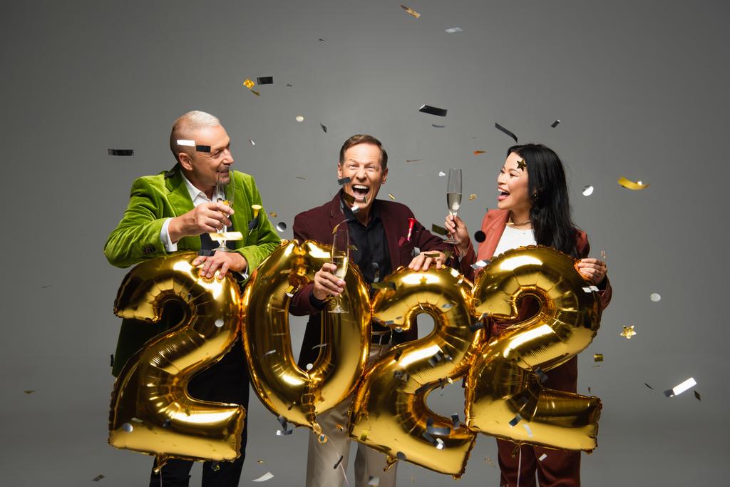 Excited interracial friends with champagne standing near balloons in shape of 2022 numbers and confetti on grey background  - Photo, Image