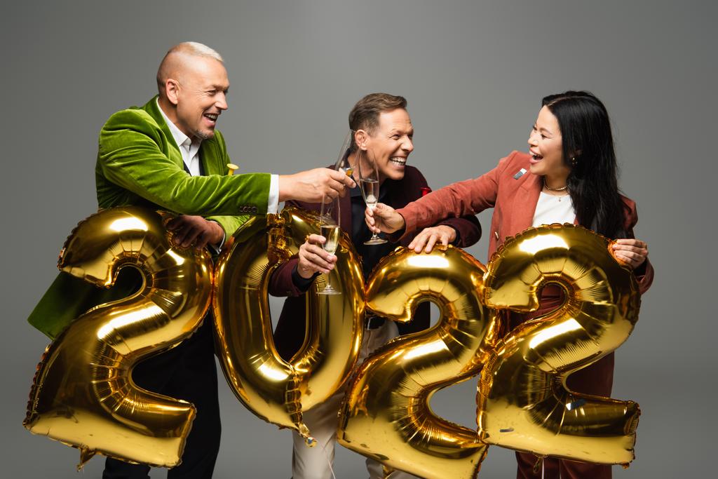 Cheerful multiethnic friends toasting with champagne near balloons in shape of 2022 numbers isolated on grey  - Photo, Image