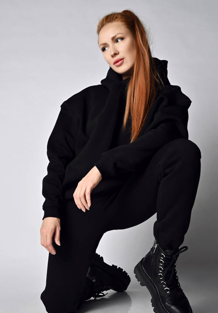 Fiatal divatos nő divatos fekete sportruházat kapucnis pulcsi, nadrág és hatalmas brutális katonai cipő áll egy térdre - Fotó, kép