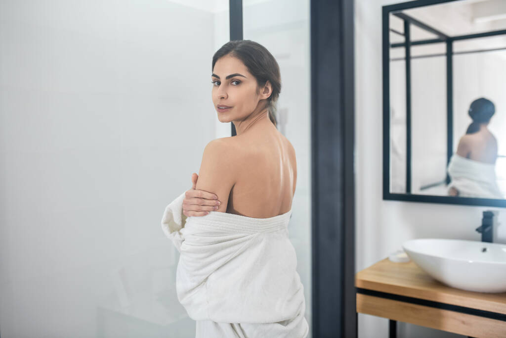 Uma jovem mulher em um manto branco com um ombro nu no banheiro - Foto, Imagem