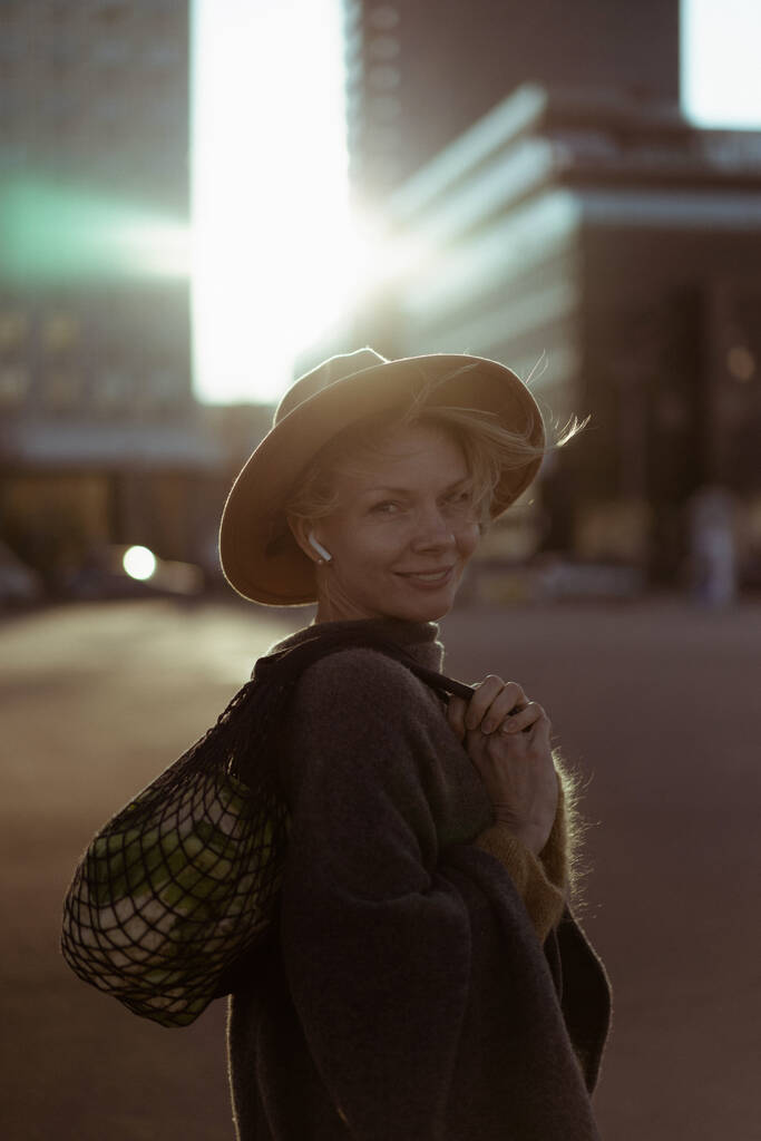 Piękna kobieta w średnim wieku w kapeluszu z krótką fryzurą w centrum dużego miasta. Portret z bliska, miękkie podświetlenie. - Zdjęcie, obraz