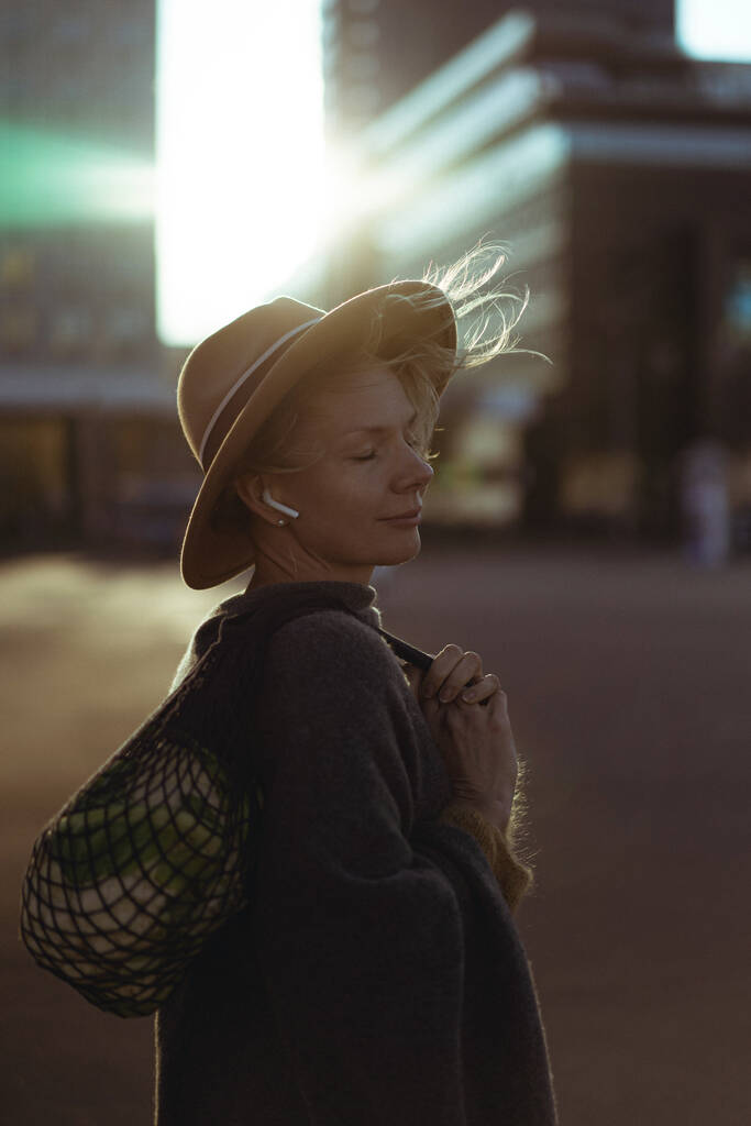 Schöne Frau mittleren Alters mit Hut und Kurzhaarschnitt im Zentrum einer Großstadt. Nahaufnahme Porträt, weiche Hintergrundbeleuchtung. - Foto, Bild