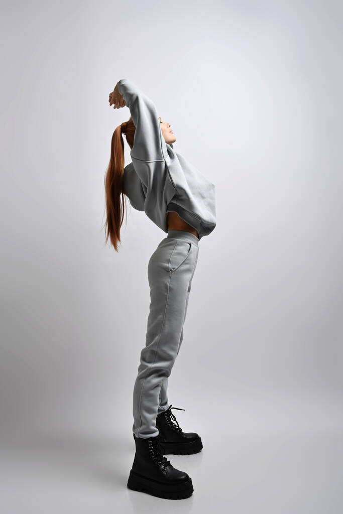 Молодая стройная спортивная женщина в спортивном костюме и массивных ботинках стоит боком, держа руки вверх, растягиваясь. Полный рост - Фото, изображение