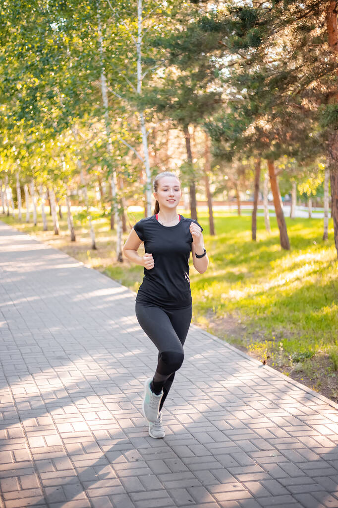 Ein Mädchen in schwarzer Sportuniform joggt im Abendpark. Selbstsportkonzept. - Foto, Bild