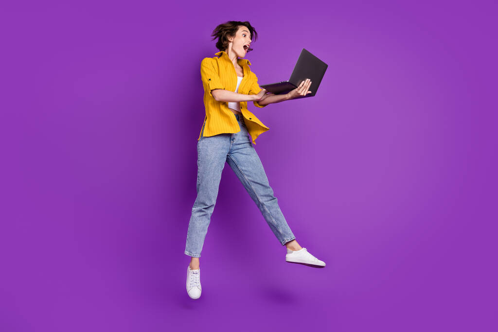 Повнометражний вигляд на тіло привабливої здивованої веселої дівчини, що стрибає за допомогою ноутбука, ізольованого на яскраво фіолетовому фіолетовому кольоровому фоні
 - Фото, зображення