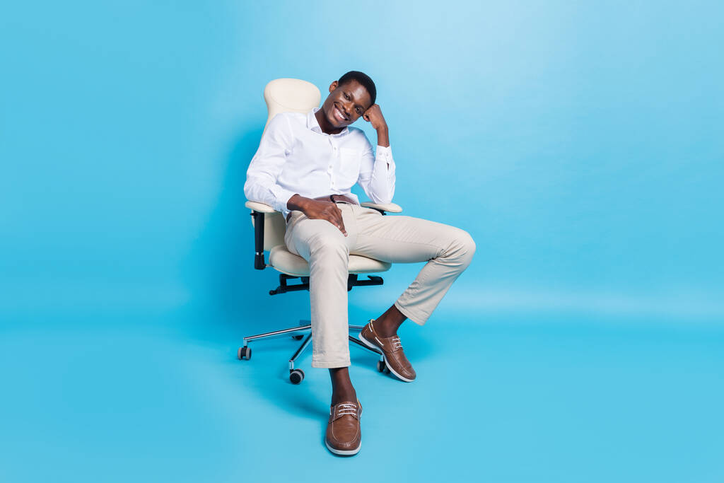 Oldukça düşünceli koyu tenli bir adamın resmi gömlekli sandalyede oturmuş izole edilmiş mavi arkaplan gülümsemesi. - Fotoğraf, Görsel