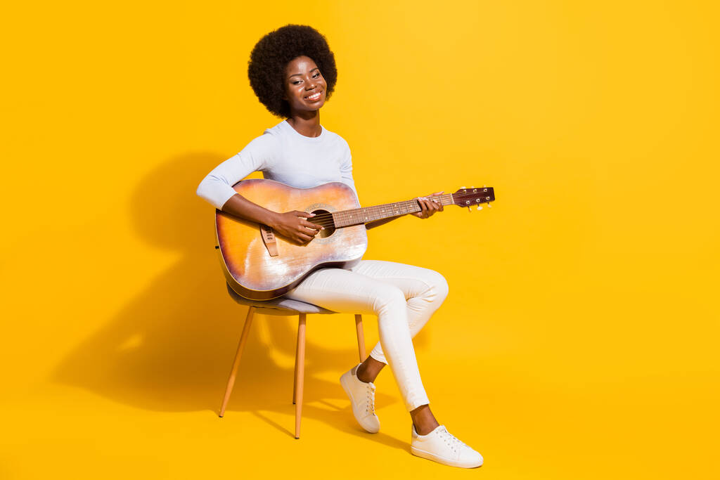 Full size profil portrét docela pozitivní tmavé kůže dáma sedí hrát na kytaru izolované na žlutém pozadí - Fotografie, Obrázek