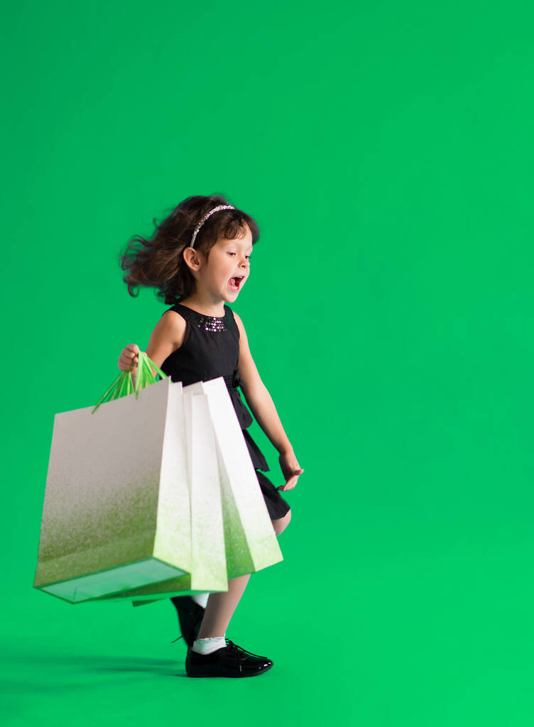 Girl in a black dress with paper bags goes shopping for discounts on a green background - Photo, Image