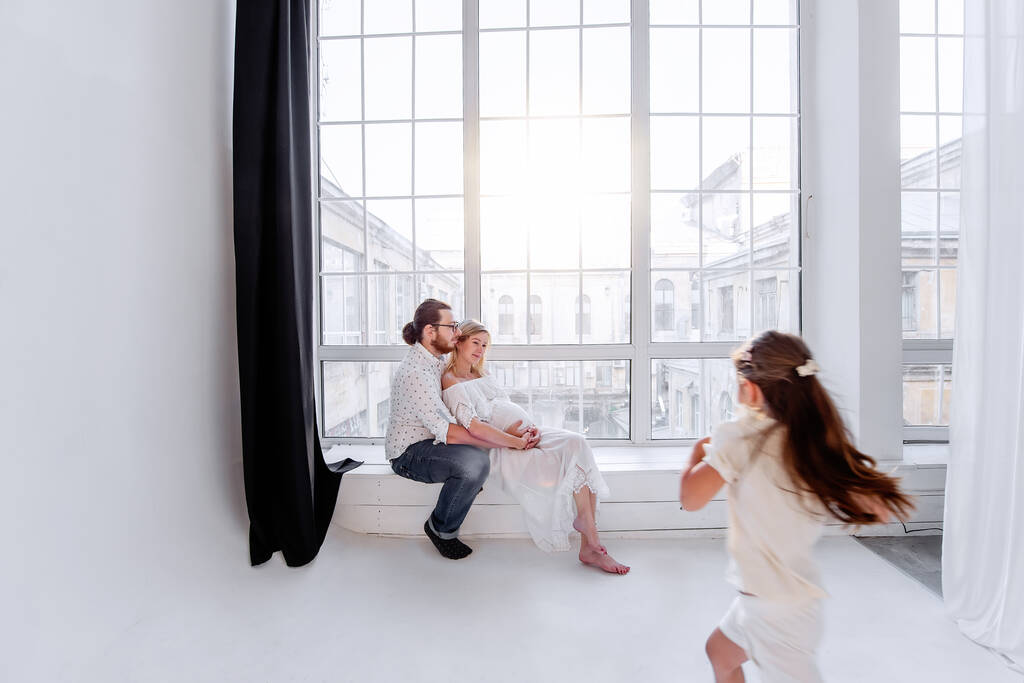 Happy family is having fun while sitting on the windowsill by the panoramic window. Young woman is pregnant, man hugs beloved. Little girl fooling around parents, dancing. Minimalist loft interior - Photo, Image