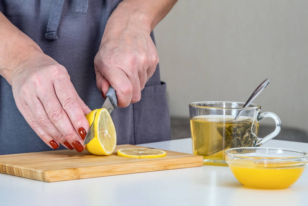 Die Gastgeberin bereitet Vitamintee zu, schneidet eine Zitronenscheibe. Tee mit Zitrone und Honig stärkt die Immunität während einer Pandemie. Schutz vor Coronavirus - Foto, Bild