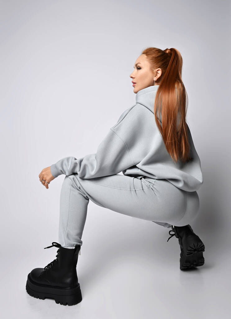 Young slim red-haired woman in sportswear hoodie, pants and black massive shoes sits squatted back to camera - Photo, Image