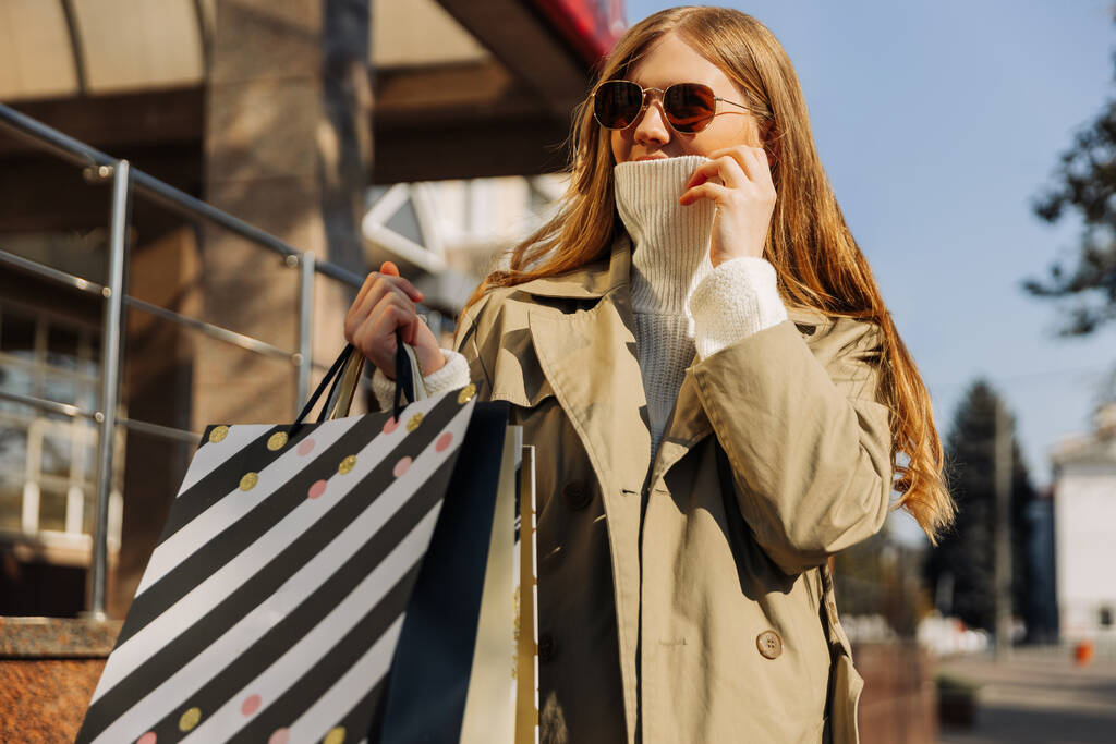όμορφη γυναίκα σε μπεζ αδιάβροχο με γυαλιά ηλίου και τσάντες για ψώνια σε λευκό πουλόβερ, καλύπτει το πρόσωπό της με φως από το κρύο. Φθινόπωρο καιρός, πώληση, υπαίθρια ψώνια - Φωτογραφία, εικόνα