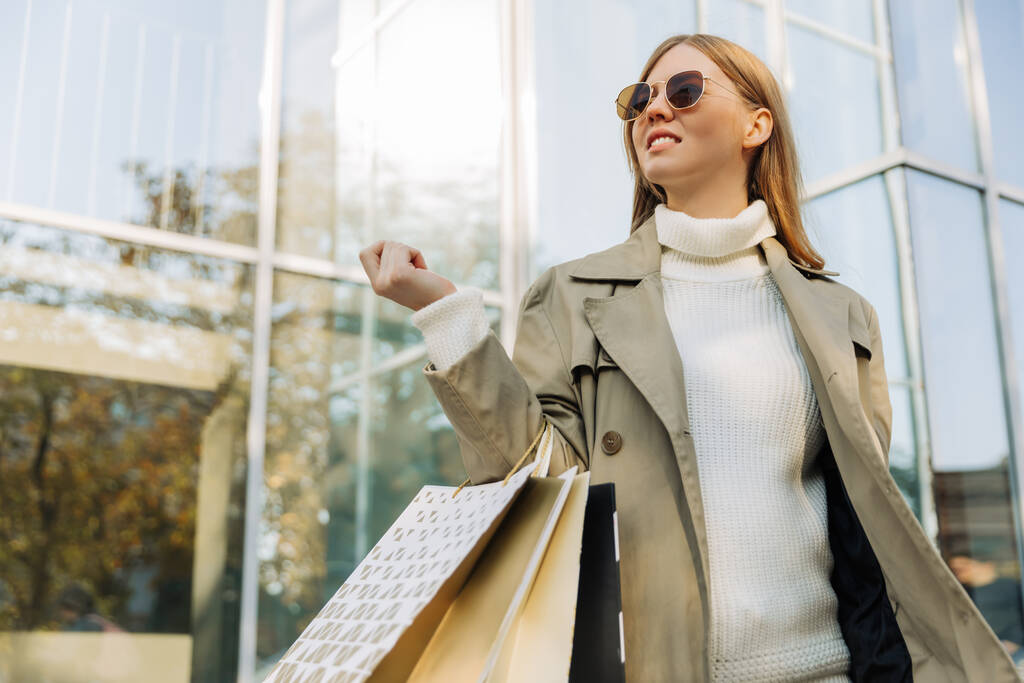 Sarışın, güneş gözlüklü, bej renkli yağmurluklu ve ellerinde alışveriş torbalarıyla bir cam binanın, bir alışveriş merkezinin önünde duruyor. - Fotoğraf, Görsel