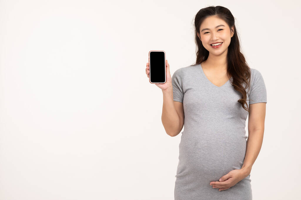 Glücklich Schwangere stehend Lächeln streichelt dicken Bauch mit Liebe halten Handy isoliert auf weißem Hintergrund, Schwangerschaft der jungen Frau genießen modernes Leben mit Smartphone, Mutterschaft Schwangerschaftskonzept - Foto, Bild