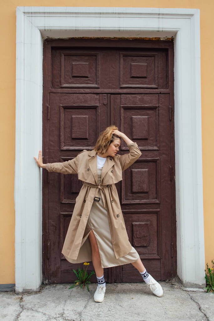 Giovane sorridente donna millenaria con i capelli selvaggi vestita con un cappotto autunnale in posa vicino alla porta di un vecchio edificio. - Foto, immagini