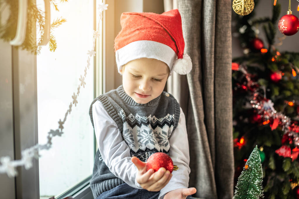 Симпатичный маленький мальчик в шляпе Санты сидит рядом с елкой в украшенной комнате и держит в руках рождественскую игрушку. Концепция подготовки к Новому году, атмосфера праздника. - Фото, изображение
