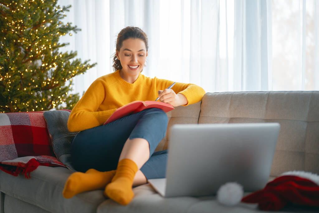 Vrouw maakt notities zittend op de bank in de woonkamer versierd voor Kerstmis. - Foto, afbeelding