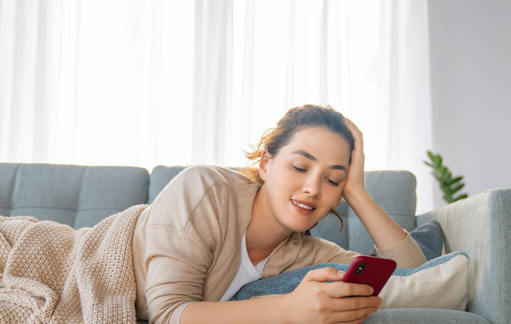 Happy casual beautiful woman is using a phone sitting on a sofa at home. - Photo, Image