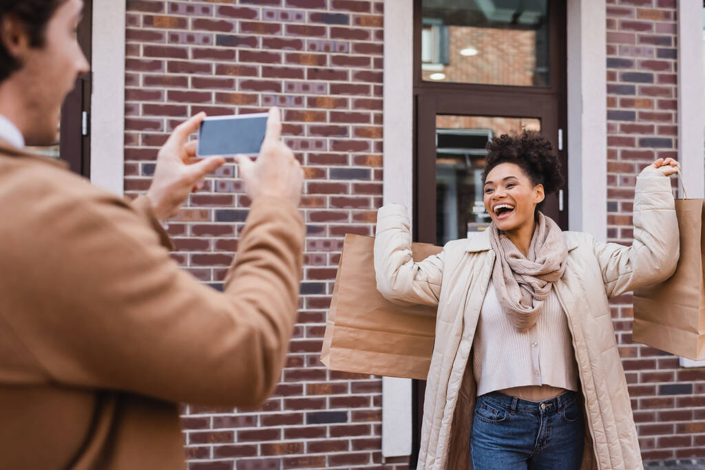 uomo sfocato scattare foto di donna afro-americana eccitato con borse della spesa - Foto, immagini