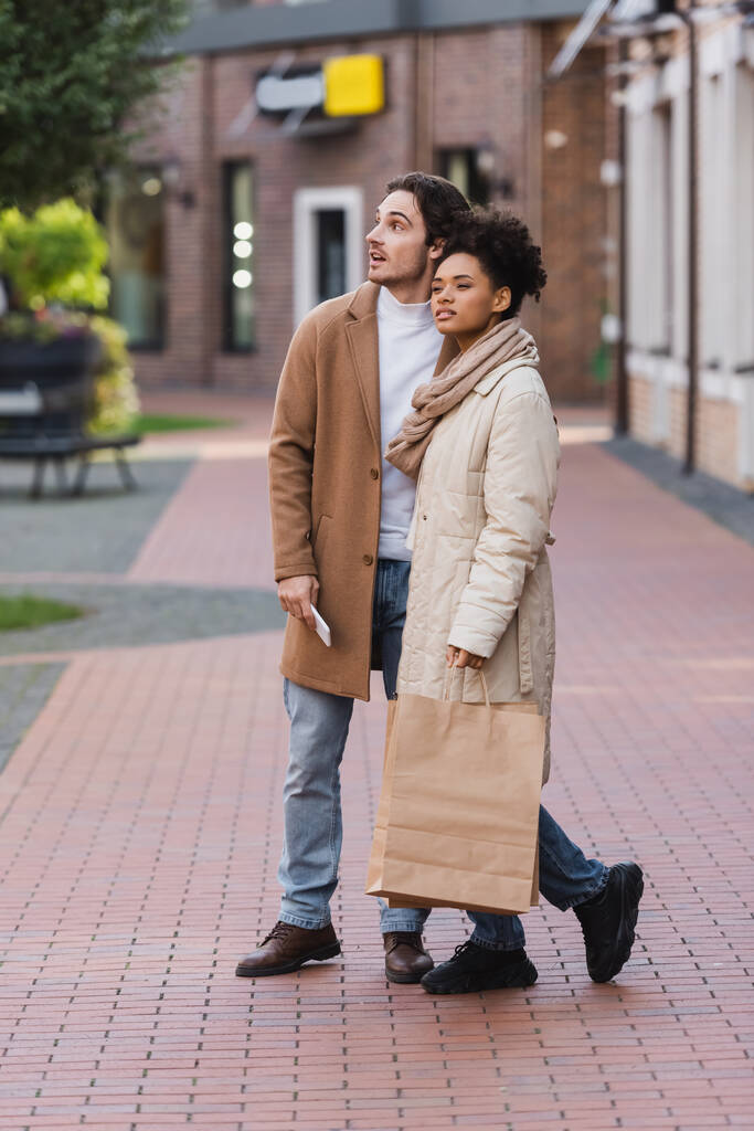 piena lunghezza dell'uomo con smartphone in piedi vicino alla ragazza afro-americana che tiene borse della spesa - Foto, immagini