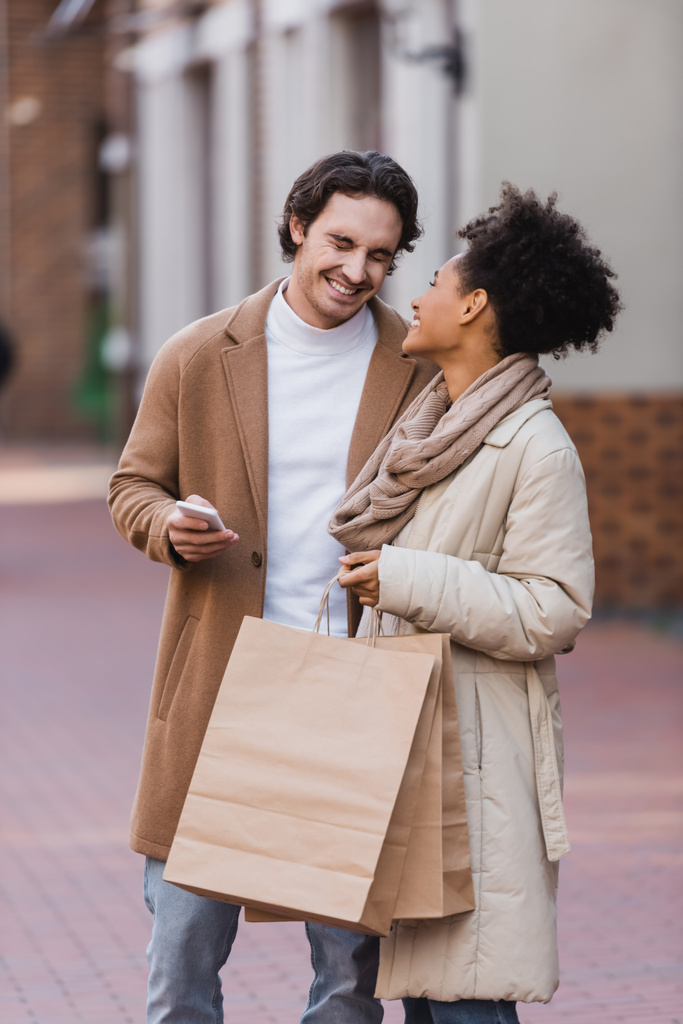 šťastný muž smartphone s úsměvem v blízkosti šťastný africký Američan žena drží nákupní tašky - Fotografie, Obrázek