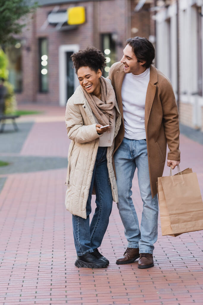 mujer afroamericana feliz con teléfono inteligente riendo cerca de novio feliz sosteniendo bolsas de compras - Foto, Imagen
