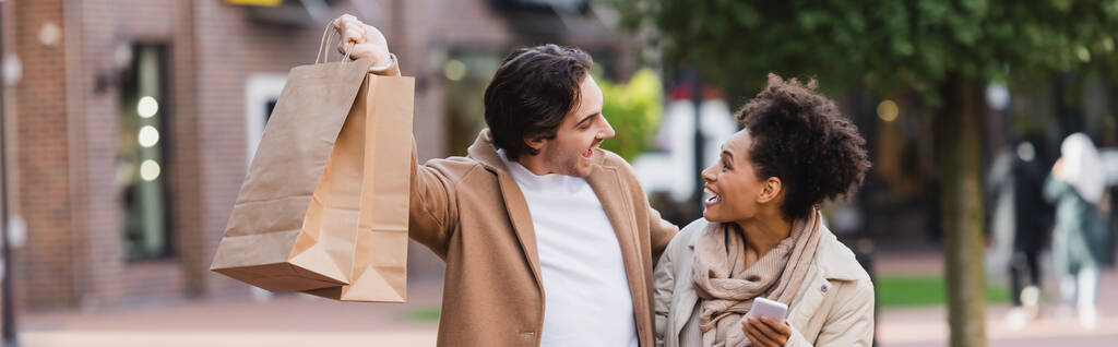 mujer afroamericana asombrada con teléfono inteligente mirando novio feliz sosteniendo bolsas de compras, pancarta - Foto, imagen