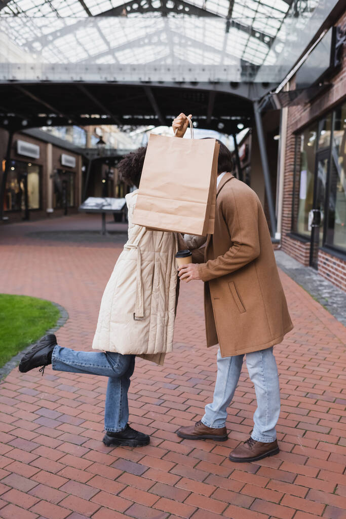 brunetka african amerykański kobieta trzyma zakupy torby podczas zakrywając twarze jej i chłopaka w centrum handlowym  - Zdjęcie, obraz
