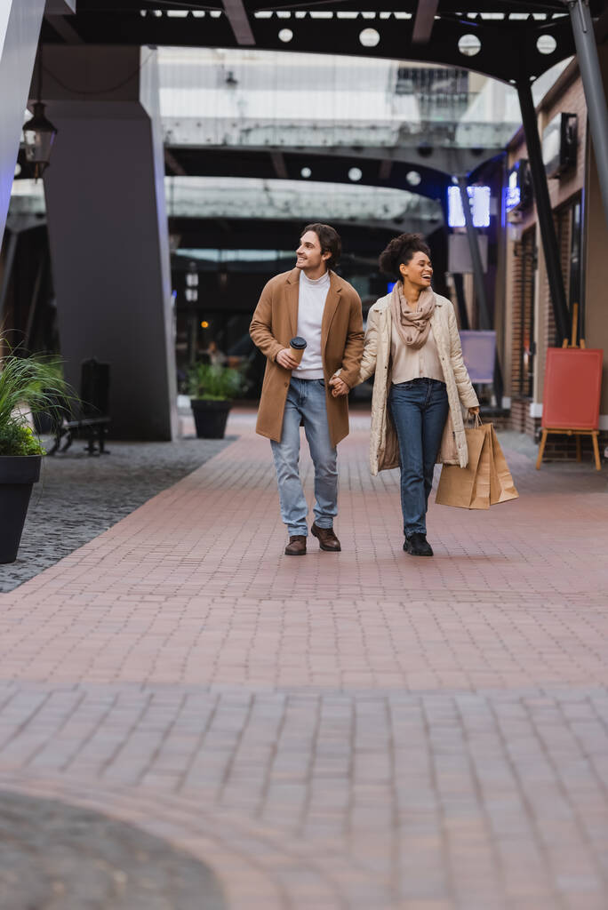 volle Länge der afrikanisch-amerikanischen Frau mit Einkaufstaschen und zu Fuß mit glücklichen Freund im Mantel in Mall  - Foto, Bild