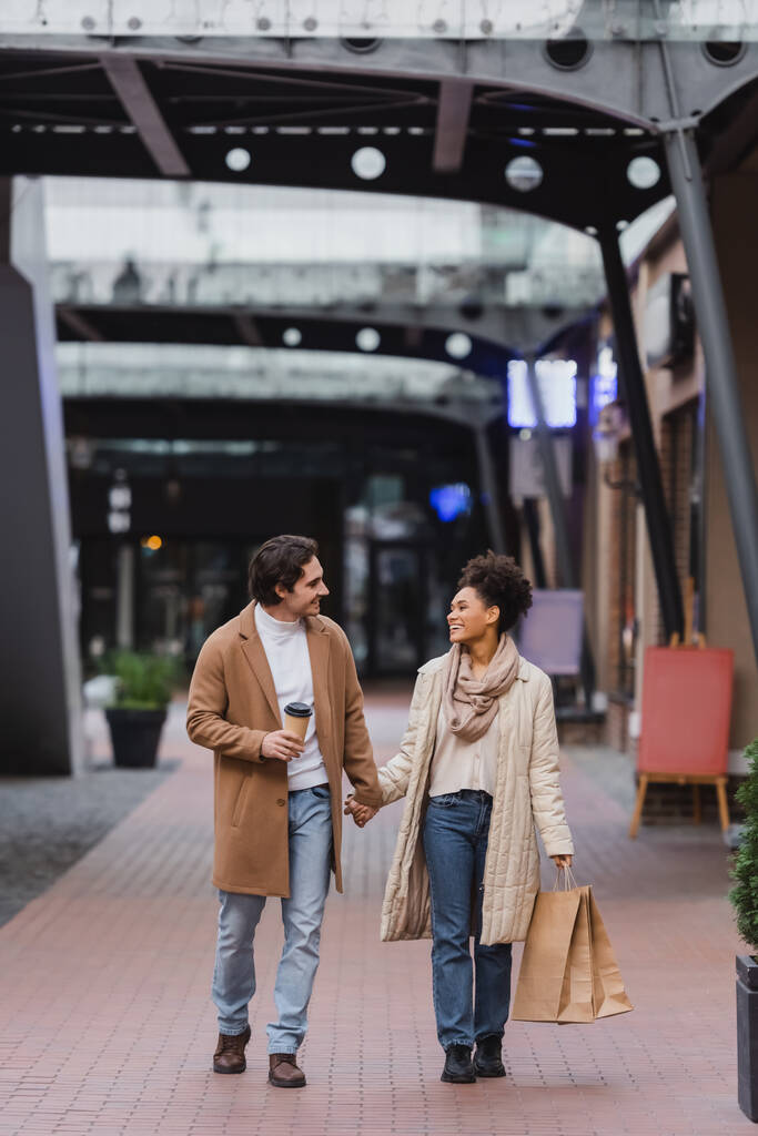 longitud completa de la mujer afroamericana sosteniendo bolsas de compras y caminando con novio alegre en abrigo en el centro comercial  - Foto, Imagen