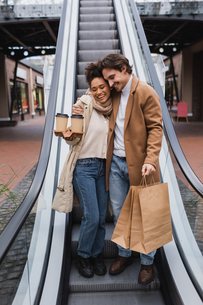 счастливая многонациональная пара с бумажными стаканчиками и сумками, обнимающая эскалатор в торговом центре  - Фото, изображение