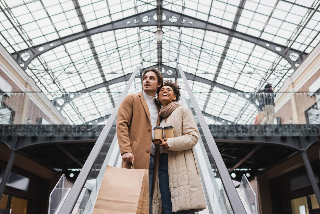 Низкий угол зрения счастливой многонациональной пары с бумажными чашками и сумками на эскалаторе в торговом центре  - Фото, изображение