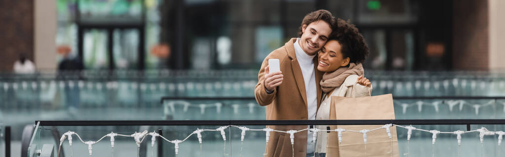 gioiosa coppia multietnica con bicchieri di carta e shopping bag scattare selfie nel centro commerciale, banner - Foto, immagini