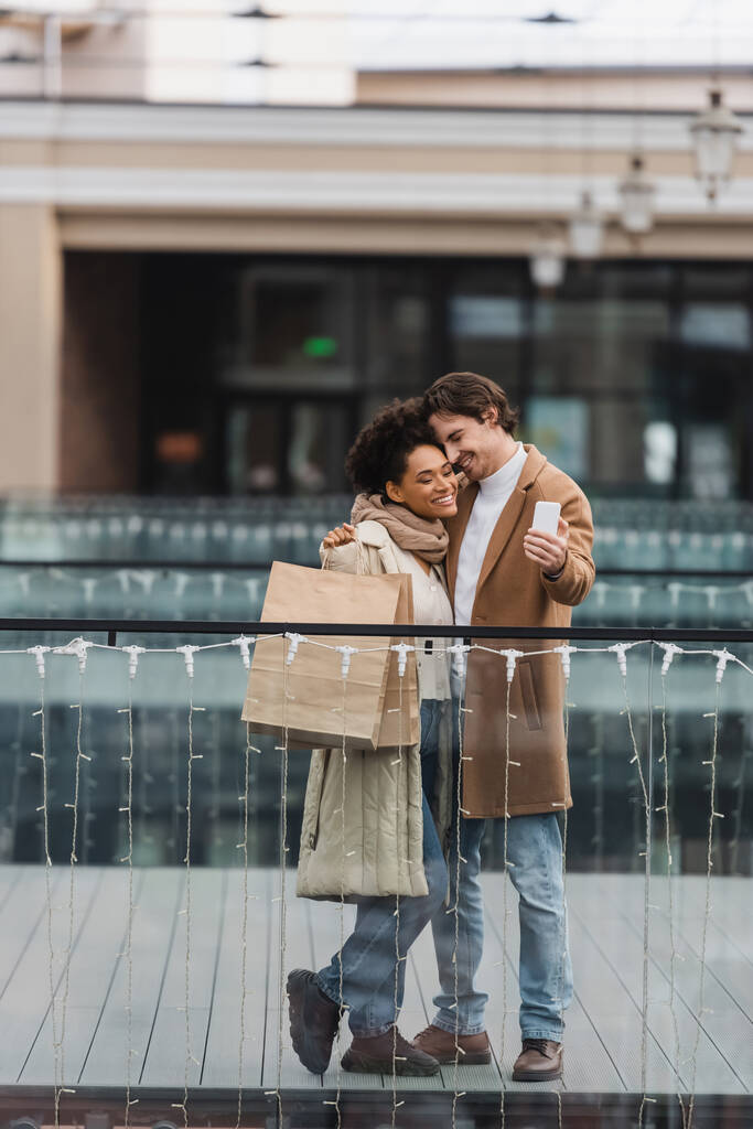 feliz pareja multiétnica con vasos de papel y bolsas de compras tomando selfie en el centro comercial  - Foto, imagen