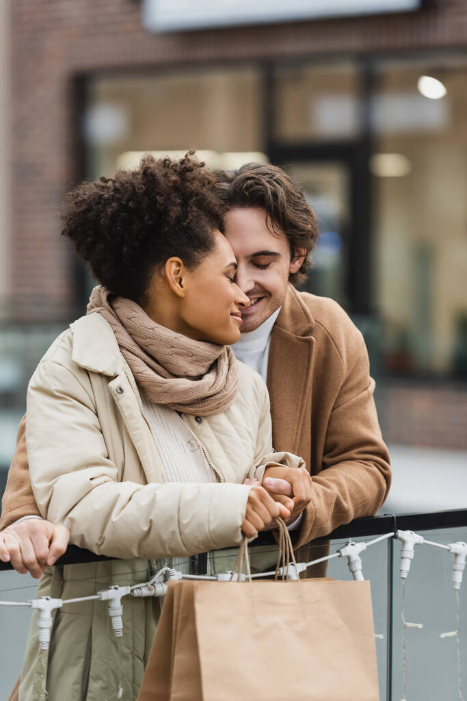 happy man holding hands with smiling african american girlfriend in shopping mall  - Photo, Image