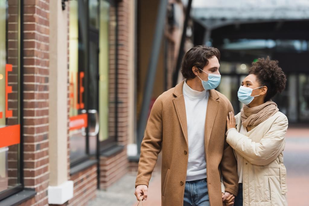 многонациональная пара в медицинских масках, держащаяся за руки во время прогулки по торговому центру  - Фото, изображение